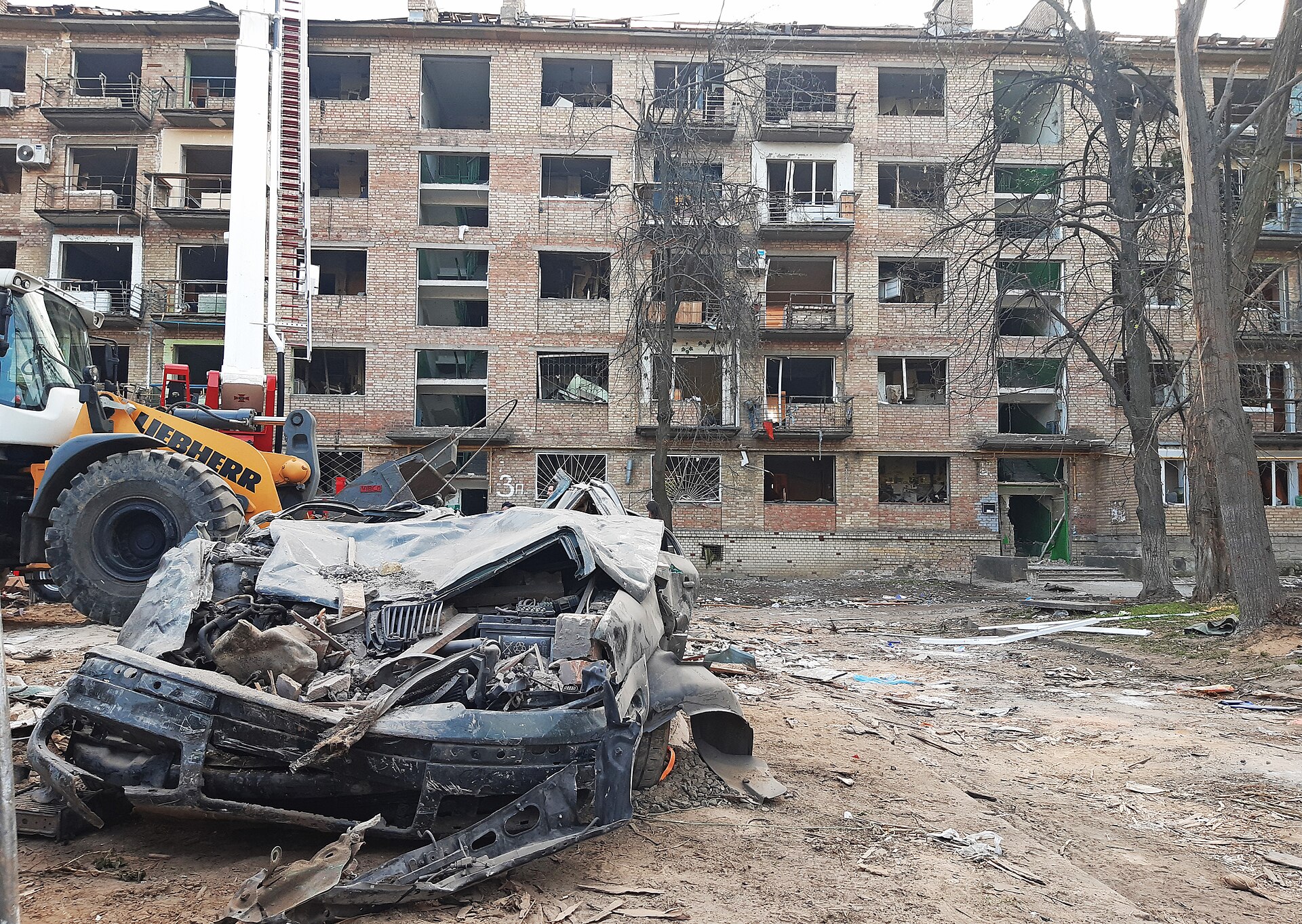 A destroyed car surrounded by rubble in front of a heavily damaged apartment building with shattered windows in Kyiv, Ukraine